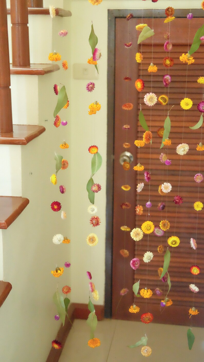 Peut inclure: Une guirlande d&eacute;corative compos&eacute;e de fleurs s&eacute;ch&eacute;es et de feuilles est suspendue au plafond. La guirlande est compos&eacute;e de petites fleurs color&eacute;es dans des tons de jaune, orange, rose et violet. Les fleurs sont entrecoup&eacute;es de feuilles vertes, ajoutant une touche naturelle &agrave; la guirlande.