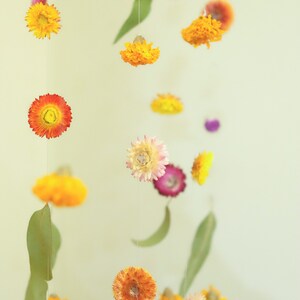 Peut inclure: Un mobile compos&eacute; de fleurs s&eacute;ch&eacute;es dans des tons de jaune, orange, rose et violet. Les fleurs sont suspendues &agrave; une ficelle blanche et dispos&eacute;es de mani&egrave;re al&eacute;atoire. Le mobile est suspendu au plafond.
