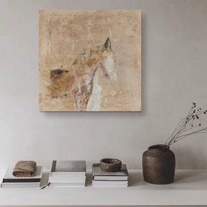 May include: A beige abstract painting of a horse's head with a textured background. The painting is framed and hanging on a white wall above a shelf with books and a vase.