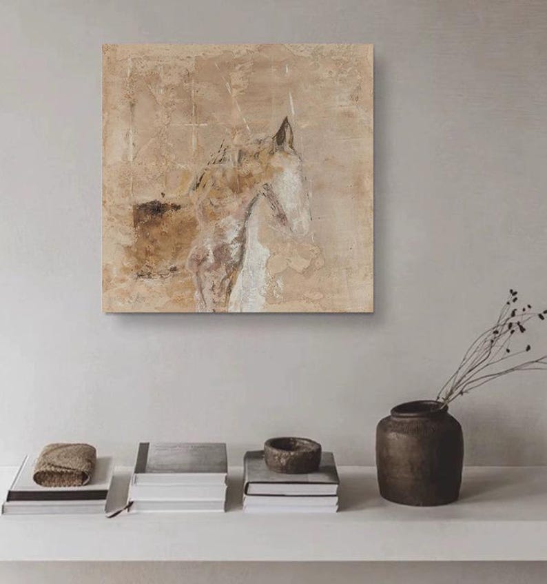 May include: A beige abstract painting of a horse's head with a textured background. The painting is framed and hanging on a white wall above a shelf with books and a vase.