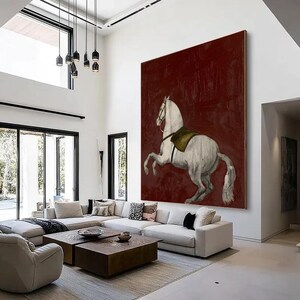 May include: Large canvas art featuring a white horse rearing up against a deep red background. The horse has a saddle and bridle. The artwork is displayed in a modern living room with white walls and furniture.