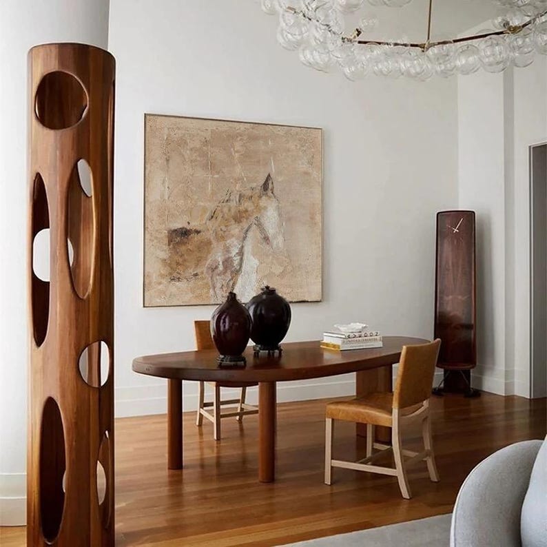 May include: A modern dining room with a wooden table and chairs. The table is oval shaped and has a light wood finish. There are two chairs with brown leather seats and a wooden frame. The room has white walls and a large abstract painting of a horse on the wall. There is a tall wooden sculpture with cutouts in the shape of circles and ovals.