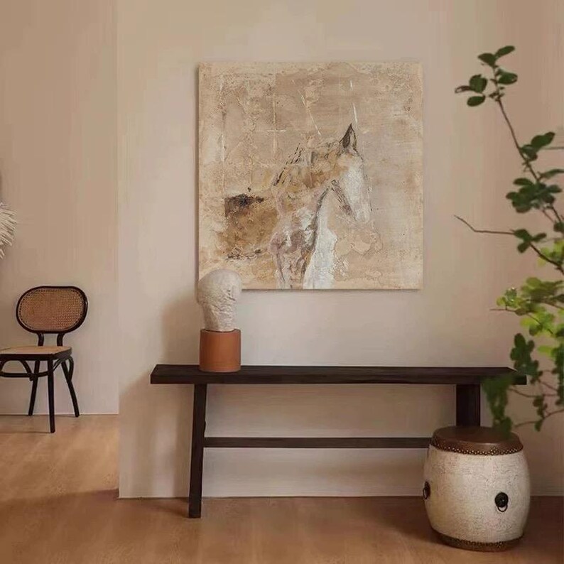 May include: A beige and brown abstract painting of a horse's head hangs on a wall above a dark wood bench. A white ceramic bust sits on a brown pedestal on the bench. A white ceramic stool with gold accents sits on the floor next to the bench.