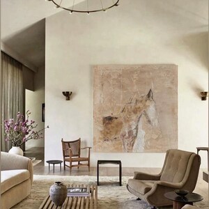 May include: A living room interior with a large abstract painting of a horse's head on the wall. The painting is in shades of brown, beige, and white. The room is decorated in a minimalist style with a beige sofa, a wooden coffee table, and two side tables. There is a brown armchair in the foreground.