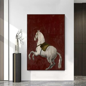 May include: A framed canvas print featuring a white horse rearing up against a deep red background. The horse has a saddle and bridle. The artwork is displayed on a white wall.