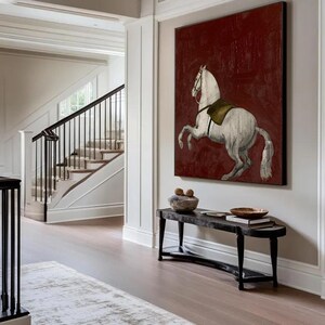 May include: A large framed canvas art piece featuring a white horse rearing up against a deep red background. The horse has a saddle and bridle. The artwork hangs above a black bench with decorative items.