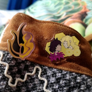 Op de afbeelding: Twee pinnen met een cartoon-koppel. De eerste pin is een geel en zwart vlamontwerp. De tweede pin toont een vrouw met zwart haar en een man met blond haar, beide met gele bloemen.