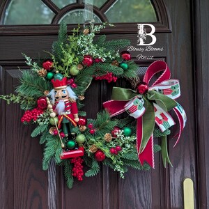 May include: A Christmas wreath with a festive design. It features a wooden nutcracker soldier, red berries, green baubles, and a large red and green bow. The wreath is displayed on a dark brown door.