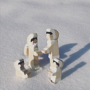 Op de afbeelding: Vier houten figuren in winterkleding, in de sneeuw. De figuren zijn lichtgekleurd met witte accenten en lijken te interageren. De scène speelt zich buiten af op een zonnige dag.