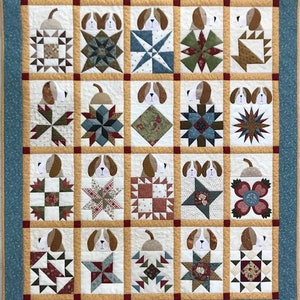 Könnte beinhalten: Eine Quiltdecke mit Patchwork-Muster in verschiedenen Mustern und Farben. Jeder Block zeigt einen Hundekopf, der hinter dem Stoff hervorlugt. Die Decke ist mit einem hellblauen Rand eingefasst.