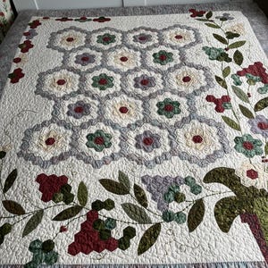 Peut inclure: Une courtepointe blanche avec un motif floral. La courtepointe présente un motif central de fleurs hexagonales dans des tons crème, verts et bordeaux. Des vignes et des bordures florales de couleurs similaires encadrent le motif central.
