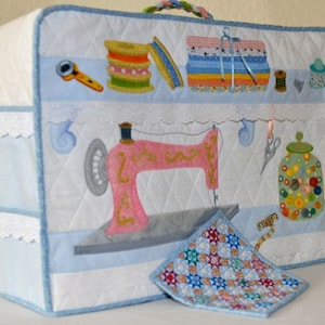 Pode incluir: Uma capa de máquina de costura acolchoada azul e branca com uma máquina de costura rosa, carretéis de linha, botões e uma fita métrica. A capa tem uma borda de renda e um pequeno quadrado acolchoado.