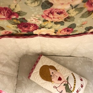 Puede incluir: Una bolsa de tela blanca con un ribete de cuadros rosa y blanco. El interior de la bolsa est&aacute; forrado con una tela blanca con un estampado floral. Una peque&ntilde;a almohada de tela blanca con un dise&ntilde;o bordado rosa y verde est&aacute; dentro de la bolsa.