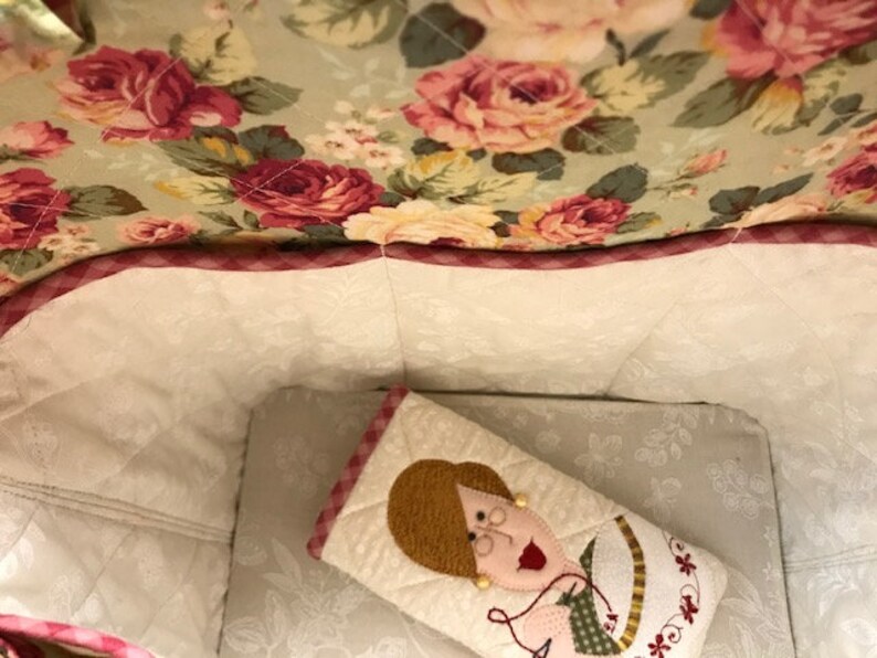 Puede incluir: Una bolsa de tela blanca con un ribete de cuadros rosa y blanco. El interior de la bolsa est&aacute; forrado con una tela blanca con un estampado floral. Una peque&ntilde;a almohada de tela blanca con un dise&ntilde;o bordado rosa y verde est&aacute; dentro de la bolsa.