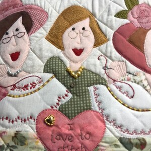 Puede incluir: Un tapiz de pared acolchado con tres mujeres que llevan sombreros y vestidos. Las mujeres sonr&iacute;en y sostienen agujas de coser. El fondo es un mosaico de tela con un coraz&oacute;n en el centro que dice "Me encanta coser".