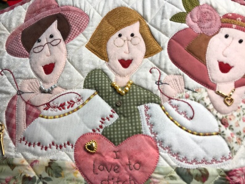 Puede incluir: Un tapiz de pared acolchado con tres mujeres que llevan sombreros y vestidos. Las mujeres sonr&iacute;en y sostienen agujas de coser. El fondo es un mosaico de tela con un coraz&oacute;n en el centro que dice "Me encanta coser".