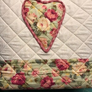 Puede incluir: Un salvamanteles de tela acolchada con un dise&ntilde;o floral en forma de coraz&oacute;n en el centro. El coraz&oacute;n est&aacute; rodeado de una tela acolchada blanca con un borde a cuadros rosa y blanco. La parte inferior del salvamanteles es un estampado floral con rosas rosas y verdes.
