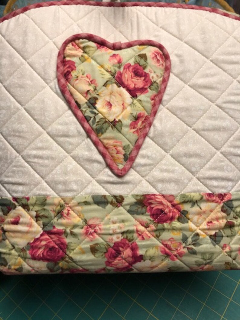 Puede incluir: Un salvamanteles de tela acolchada con un dise&ntilde;o floral en forma de coraz&oacute;n en el centro. El coraz&oacute;n est&aacute; rodeado de una tela acolchada blanca con un borde a cuadros rosa y blanco. La parte inferior del salvamanteles es un estampado floral con rosas rosas y verdes.