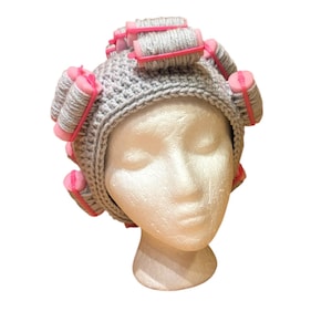 May include: A gray crocheted hat with pink and gray hair rollers. The hat is on a white mannequin head. The hair rollers are evenly spaced around the hat, creating a unique and playful design. The hat is a fun accessory.