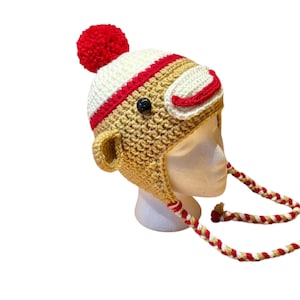 May include: A handmade crochet hat in the shape of a monkey, featuring a red pom-pom, ear flaps, and braided ties. The hat is primarily tan with white and red accents, and a black button eye.