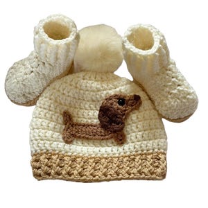 A cream crocheted baby hat with a brown dachshund applique and a cream pom pom on top. The hat is paired with matching cream crocheted booties.