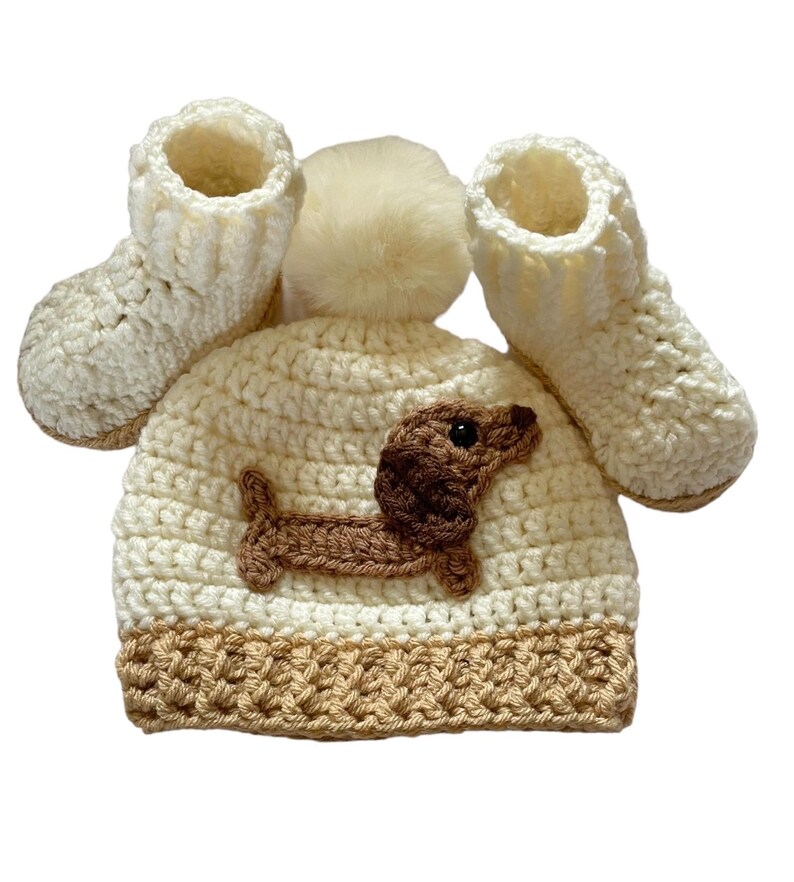 A cream crocheted baby hat with a brown dachshund applique and a cream pom pom on top. The hat is paired with matching cream crocheted booties.