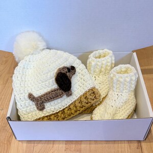 Sausage dog baby gift set with  handmade crochet hat and matching booties - a neutral baby shower present. photo shows a Close up of dachshund applique on baby hat with soft yarn texture and fluffy pom pom top