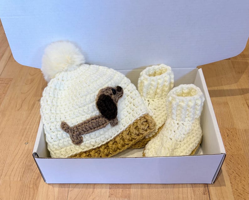 Sausage dog baby gift set with  handmade crochet hat and matching booties - a neutral baby shower present. photo shows a Close up of dachshund applique on baby hat with soft yarn texture and fluffy pom pom top