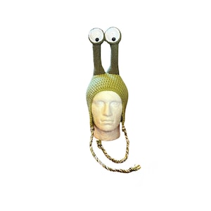 May include: A light green crocheted hat featuring two tall, stalk-like protrusions topped with large, circular white eyes with black pupils. The hat includes two braided green and white cords hanging from the sides, designed to tie under the chin.