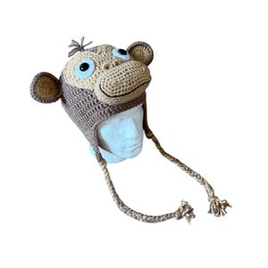 May include: A brown and tan crocheted monkey hat featuring a protruding muzzle, two round ears, and a small tuft of hair on top. The hat includes two long, braided earflaps with tasseled ends and two large, round black and white plastic eyes.