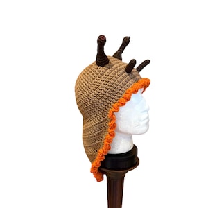May include: A tan crochet hat featuring a textured surface and a bright orange ruffled trim along the edge. The hat includes four dark brown, stalk-like protrusions extending from the top, with two of the stalks featuring small red eye details.