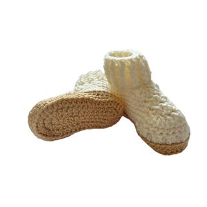 A pair of crocheted baby booties in cream and beige. The booties have a textured design and are made with soft yarn.