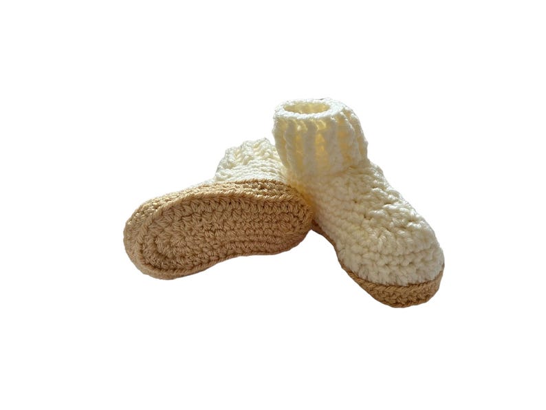 A pair of crocheted baby booties in cream and beige. The booties have a textured design and are made with soft yarn.
