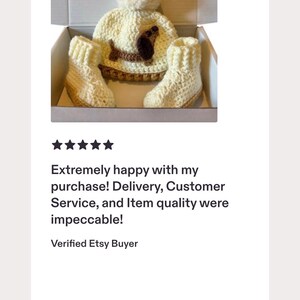 Peut inclure: Un bonnet en crochet couleur cr&egrave;me avec un gros pompon et une application de teckel marron, accompagn&eacute; d'une paire de chaussons en crochet assortis de couleur cr&egrave;me. L'ensemble est pr&eacute;sent&eacute; dans une bo&icirc;te en carton blanc ouverte.