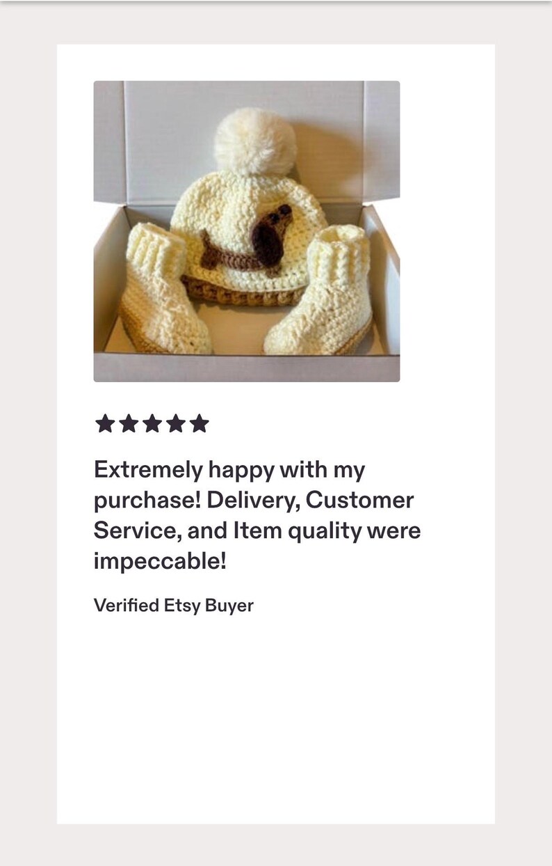 Peut inclure: Un bonnet en crochet couleur cr&egrave;me avec un gros pompon et une application de teckel marron, accompagn&eacute; d'une paire de chaussons en crochet assortis de couleur cr&egrave;me. L'ensemble est pr&eacute;sent&eacute; dans une bo&icirc;te en carton blanc ouverte.