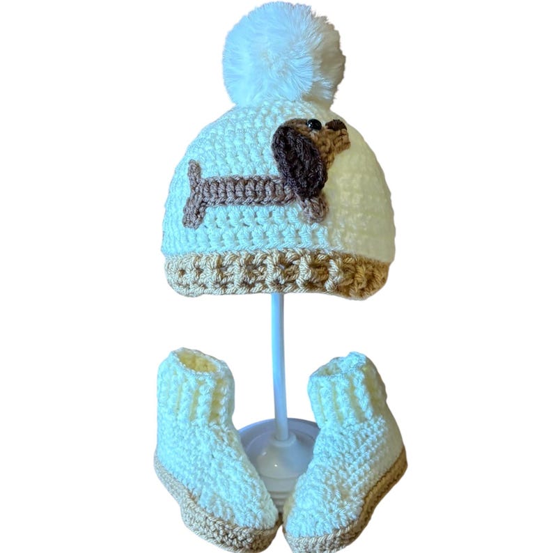 May include: A white crocheted beanie with a large white pom-pom on top and a tan brim. A brown crocheted dachshund dog applique is centered on the front of the beanie. A pair of matching white crocheted booties with tan soles completes the set.