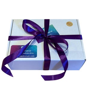 image shows a white gift box tied with a purple satin ribbon to show how baby hat and booties sets are gift wrapped free of charge. The package has a brown paper sticker with the text "Handmade with love" and a small heart.