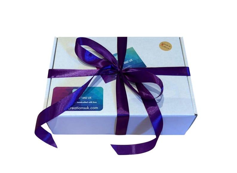 image shows a white gift box tied with a purple satin ribbon to show how baby hat and booties sets are gift wrapped free of charge. The package has a brown paper sticker with the text "Handmade with love" and a small heart.