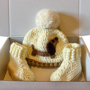 May include: A cream-colored crocheted beanie with a large matching pom-pom on top and a brown dachshund appliqu&eacute; on the front, accompanied by a pair of matching cream-colored crocheted booties with brown soles, all presented in a white box.