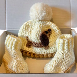 May include: A cream-colored crocheted beanie with a matching pom-pom and a brown dachshund appliqu&eacute;, accompanied by two cream-colored crocheted booties with tan trim, all presented together in a white box.