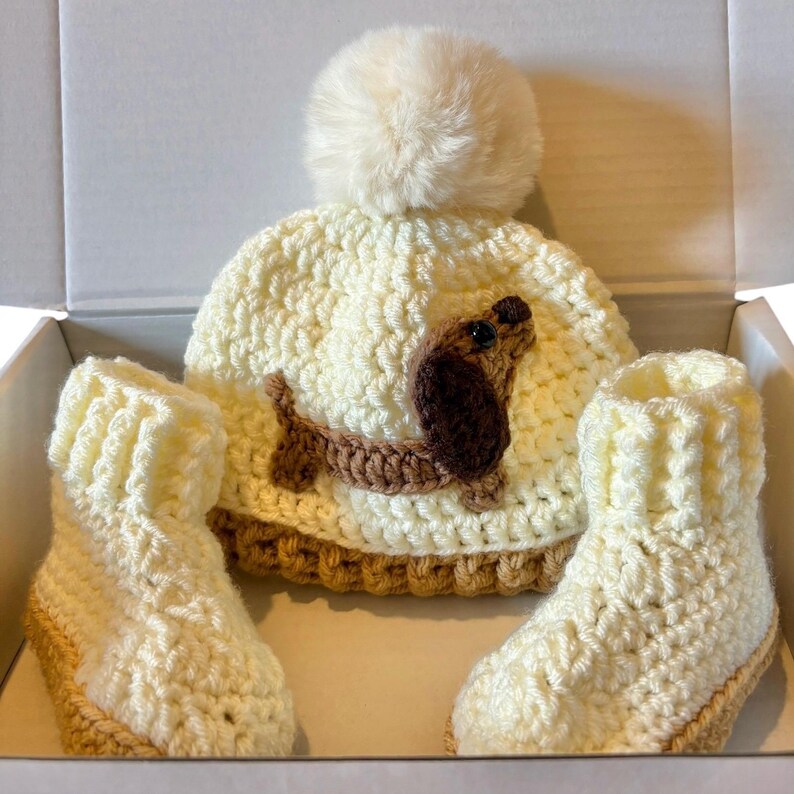 May include: A cream-colored crocheted beanie with a matching pom-pom and a brown dachshund appliqu&eacute;, accompanied by two cream-colored crocheted booties with tan trim, all presented together in a white box.