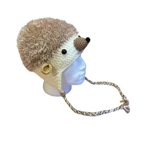 May include: A crochet hat designed to resemble a hedgehog, featuring a textured tan faux-fur top, a cream-colored base, a small brown pointed nose, black button eyes, a round ear, and two long braided ties hanging from the sides of the headpiece.