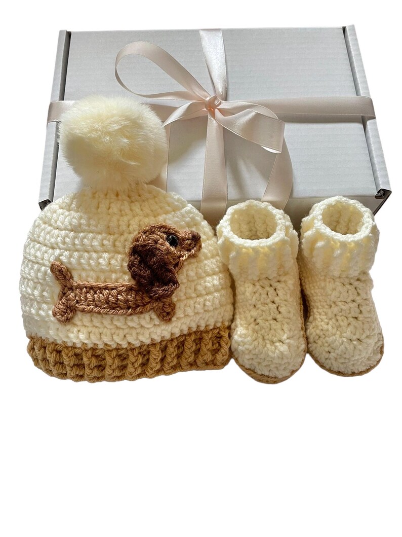 A cream and brown crocheted baby hat with a brown dachshund applique and a white pom pom on top. The hat is in a white gift box with a satin ribbon. There are also two cream crocheted booties in the box.