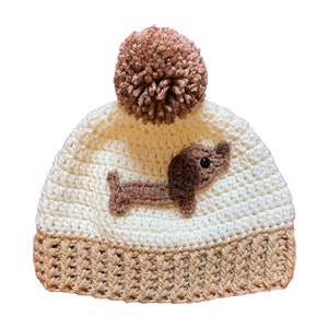 May include: White and brown crocheted beanie with a brown dachshund applique and a brown pom pom on top.