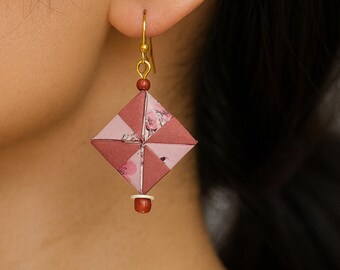 May include: A close-up of a single earring. The earring features a geometric, diamond-shaped design in shades of pink and red, with floral accents. It hangs from a gold-coloured hook and is adorned with small beads.