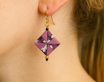 May include: A close-up of a single earring. The earring has a square, origami-style design in shades of purple with a floral pattern. It hangs from a gold-coloured hook and has a small bead at the bottom. The earring is displayed on a person's ear.