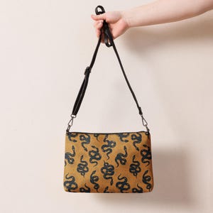 May include: A mustard yellow crossbody bag with a black adjustable strap. The bag has a snake print pattern in black and white.