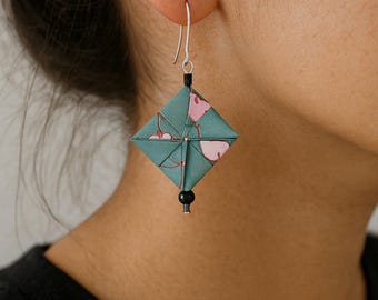 May include: A single origami earring with a square, diamond-shaped design. The earring is teal with pink floral accents and a black bead at the bottom. The earring hangs from a silver hook.