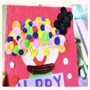 Pu&ograve; includere: Un'artigianato in feltro colorato che rappresenta un viso sorridente con un'ampia acconciatura di pompon colorati e le parole "Happy Day" su uno sfondo viola.
