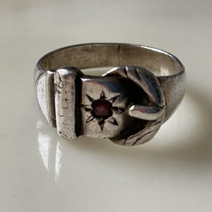 May include: A silver-toned metal ring designed to resemble a buckled belt. The band features a buckle mechanism with a central star-shaped engraving containing a small, dark red circular stone. The metal surface shows a polished, reflective finish.